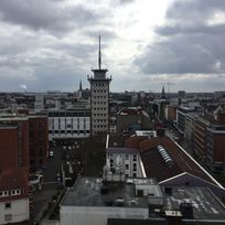 Zu sehen ist der Ausblick auf eine Stadt mit vielen Häusern und spitzen Türmen. Links im Bild ist ein freier Platz zu sehen, auf dem Autos parken. Rechts befindet sich eine Straße zwischen den Häusern, auf der Autos fahren. In der Mitte des Bildes ragt ein spitzer Turm zwischen den Häusern empor. Der Himmel ist blau und bewölkt.