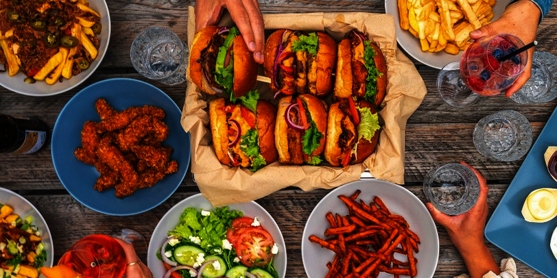 Ein Tisch voll mit Burgern und Pommes, am Bildrand einige Hände, die nach dem Essen greifen