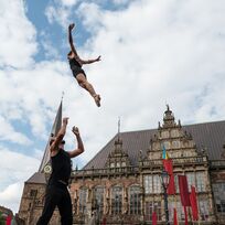 Zwei Akrobat*innen führen auf dem Bremer Marktplatz ein Kunststück auf. Im Hintergrund ist das Rathaus zu sehen. Der Himmel ist blau mit ein paar Wolken.