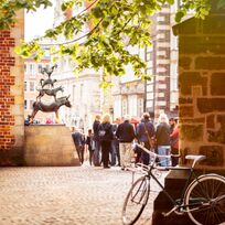 Die Stadtmusikanten Statue ist umringt von Menschen, im Vordergrund steht ein Fahrrad an die Wand gelehnt und Äste mit grünen Blättern hängen ins Bild herein.