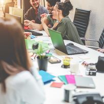 Junge, gut gelaunte Leute sitzen an einem großen Besprechungstisch und tauschen sich aus. Auf dem Tisch stehen Laptops und andere Arbeitsutensilien.