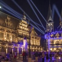 Der beleuchtete Bremer Dom und das beleuchtete Bremer Rathaus bei Nacht. Davor strahlen zahlreiche Scheinwerfer in den Himmel.
