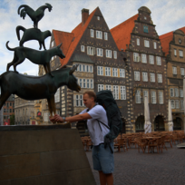 Ein Mann mit großem Reiserucksack steht vor der Statue der Bremer Stadtmusikanten und umfasst die Beine des Esels mit beiden Händen.