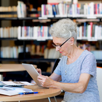 Eine ältere Dame sitzt mit ihrem iPad in der Bibliothek und studiert