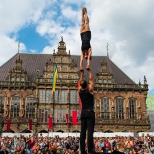 Zwei Akrobat*innen führen auf dem Bremer Marktplatz ein Kunststück auf. Im Hintergrund ist das Rathaus und Publikum zu sehen. Der Himmel ist blau mit ein paar Wolken.