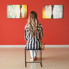 Eine Frau sitzt mit dem Rücken zur Kamera vor einer Wand, an der zwei Bilder aufgehängt sind