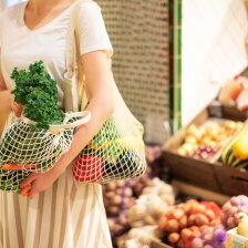 Eine Person hat zwei grob gewebte Einkaufstaschen mit Obst und Gemüse bei sich. Einen hat sie auf der Schulter liegen und den anderen in der Hand. Im Hintergrund befindet sich weiteres Obst und Gemüse.