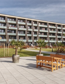 Außenansicht eines großen Wohnhauses mit Balkonen. Davor ein Platz mit Holzbänken und Palmen in Blumentöpfen. 