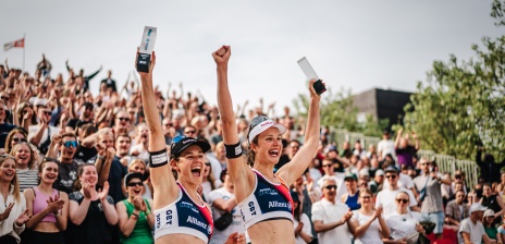 Two women cheer and hold up a trophy. Grandstand in the background.