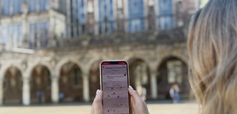 Someone holds their cell phone with the DBG app in front of the town hall and looks at the map “UNESCO World Heritage Town Hall and Roland.”