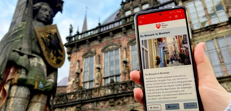 Jemand hält ein Smartphone vor dem Rathaus in Bremen. Auf dem Bildschirm sieht man die Startseite von "Dein Bremen Guide".
