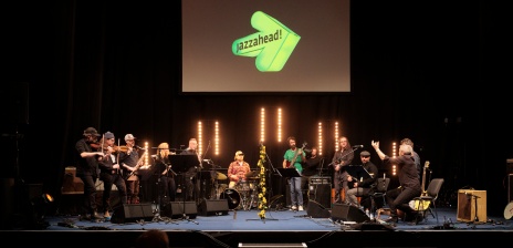 Bühne mit vielen Jazzmusiker*innen, die Musik spielen. Im Hintergrund wird das Logo der jazzahead an die Wand geworfen.
