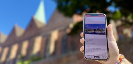 Eine Hand hält ein Handy auf welchem man Informationen über die St. Martini Kirche durch den DBG erhält. Im Hintergrund befindet sich die St. Martini Kirche.