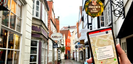 Auf einem Smartphone wird auf einer Karte das Restaurant "Beck's in 'n Snoor" angezeigt. Im Hintergrund sieht man das Restaurant.