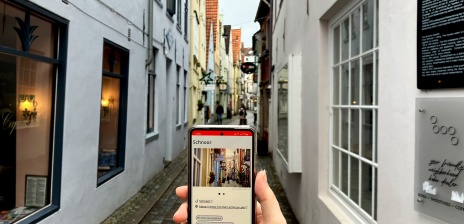 Im Schnoor hält jemand ein Smartphone. Darauf sieht man Informationen zum Schnoor in "Dein Bremen Guide".