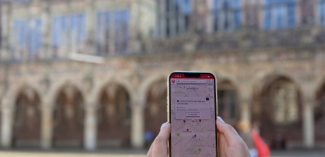 Jemand hält sein Handy mit der DBG App vor das Rathaus und schaut sich auf der Karte "UNSECO-Welterbe Rathaus und Roland" an. 