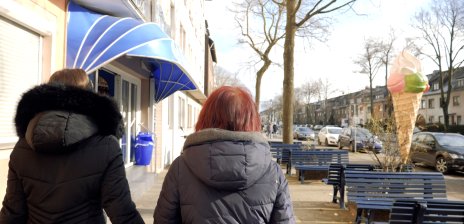 Zu sehen sind zwei weibliche Personen von hinten, die beide eine Winterjacke tragen und an einer Straße entlanggehen. Rechts stehen blaue Bänke und eine große Eiswaffel-Figur. Links ist eine blaue Markise zu sehen.