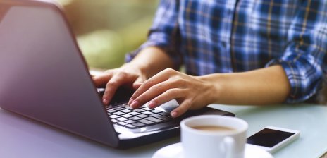 Eine Frau tippt auf einer Laptoptastatur, daneben Kaffeetasse und Smartphone.