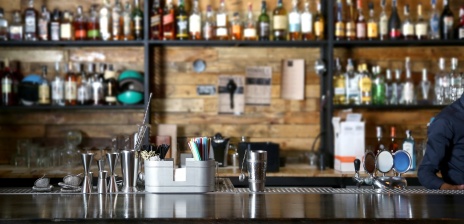 Eine Theke ist im Zentrum des Bildes. Am Rand steht ein Barkeeper. Im Regal sind verschiedene Flaschen und Utensilien zum Cocktails mixen. 