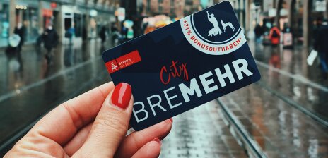 Eine Hand mit rot lackierten Fingernägeln hält den "BreMEHR"-Gutschein vor der Kulisse der Bremer Innenstadt. 