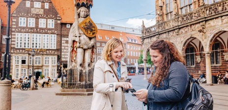 Zwei Frauen gucken sich auf dem Bremer Marktplatz den Audioguide an und haben Kopfhörer im Ohr.