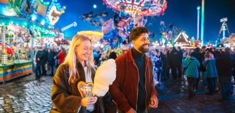 Zwei lächelnde Menschen laufen an den bunt beleuchteten Fahrgeschäften und Ständen des Freimarkts entlang.