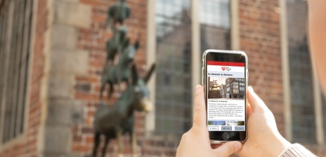 Jemand hält ein Smartphone vor den Stadtmusikanten. Auf dem Bildschirm sieht man die Startseite von "Dein Bremen Guide".