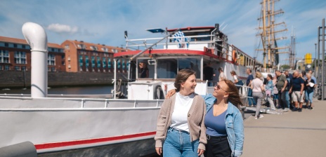 Zwei Frauen lachen sich an. Im Hintergrund gehen Leute an Bord eines Schiffes.