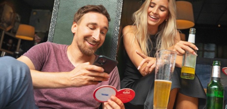 Zwei Personen trinken zusammen was. Die eine Person macht ein Foto mit dem Handy von einem Bierdeckel.