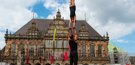 Zwei Akrobat*innen führen auf dem Bremer Marktplatz ein Kunststück auf. Im Hintergrund ist das Rathaus und Publikum zu sehen. Der Himmel ist blau mit ein paar Wolken.