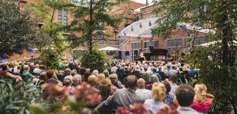 Publikum des Musikfests sitzt vor einer Open-Air-Bühne zwischen grünen Bäumen und historischen Gebäuden.