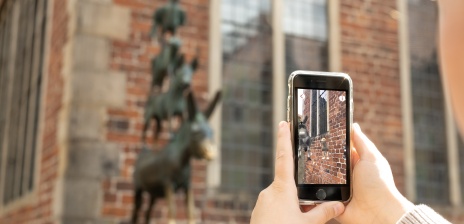 A smartphone with animated Town Musicians figures is held in front of the statue of the Town Musicians.