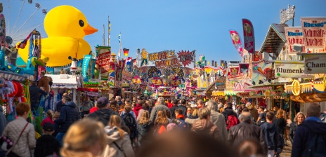 Fairground stalls and carousels on the Easter meadow. Visitors stroll along the middle of the stalls. 