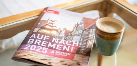 Ein Bremer Reisekatalog 2025 liegt auf einem Glastisch, eine Tasse Kaffee steht daneben.