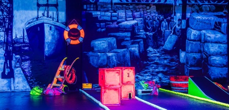 Ein mit Schwarzlicht ausgeleuchteter Innenraum, in dem sich verschiedene Minigolfbahnen befinden. 