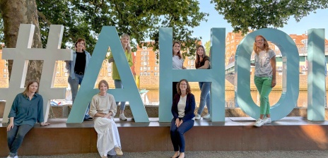 Acht Frauen stehen verteilt vor, hinter und zwischen großen Moin-Buchstaben und lächeln in die Kamera.