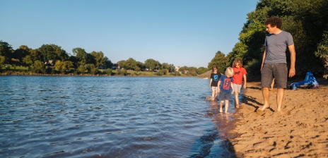 Ein Mann läuft mit Kindern am Weserstrand entlang. Die Kinder kühlen ihre Füße im Wasser ab. 