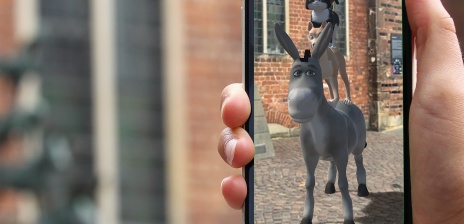Im Vordergrund sind die Bremer Stadtmusikanten in 3D durch ein Smartphone zu sehen. Im Hintergrund ist die Statue der Stadtmusikanten zu sehen. 