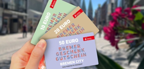 Drei Geschenkgutscheine werden vor der Kulisse der Bremer Einkaufsstraße Obernstraße in die Luft gehalten.