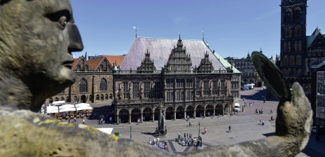 Blick auf ein steinernes, historisches Gebäude, welches auf einem Platz steht, auf dem sich mehrere Menschen aufhalten. Zudem zeigt die Aufnahme einen Turm des Bremer Doms. 