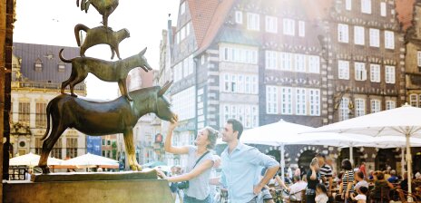 A woman and a man stand in front of the Bremen Town Musicians. The woman touches the donkey's mouth and smiles. The man stands behind her and watches the statue. 