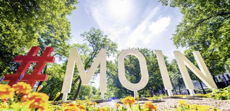 Der Schriftzug "Moin" in den Wallanlagen. Im Vordergrund sind zahlreiche bunte Blumen zu sehen. 