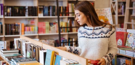 Eine junge Frau steht in einer Buchhandlung vor einem tischhohen Regal und schaut sich die Bücher an. Im Hintergrund stehen große Bücherregale.