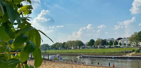 Ausblick auf den Strand am Café Sand, auf der Weser die Sielwallfähre und ein Binnenschiff, am Strand Menschen in Badekleidung, am linken Bildrand grüne Blätter eines Baumes