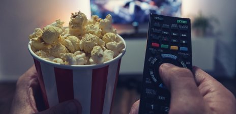 Eine Person sitzt vor einem Fernseher und hält eine Fernbedienung und einen Becher Popcorn in der Hand