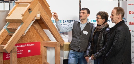 Zwei Männer und eine Frau stehen an einer Holzhütte und begutachten das Material. 