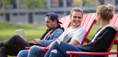 Drei Studierende, eine blone Frau mit Dutt, ein blonder Mann und ein brünetter Mann mit Baseballkappe, sitzen auf roten Holzstühlen auf dem Campus der Universität Bremen. Sie reden miteinander und lachen. Der Mann mit der Kappe schaut auf seinen Laptop.