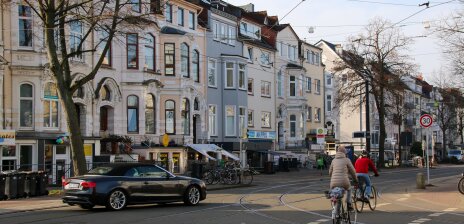 Zwei Fahrradfahrer und ein Auto fahren über eine Kreuzung. Im Hintergrund reihen sich Altbremer Häuser aneinander. 
