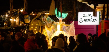 Winter market visitors stand together. Dressed up stilt walkers stand between them.