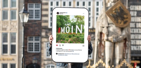 In der Mitte des Bildes wird eine Art Smartphone-Bildschirm von zwei Händen hochgehalten - darauf zu seine ist ein Social Media Post, der ein Foto eines MOIN-Schriftzuges auf einer Blumenwiese zeigt. Hierbei handelt es sich um ein Exponat einer Ausstellung. Im Hintergrund ist unscharf der Bremer Marktplatz mit Roland zu sehen.